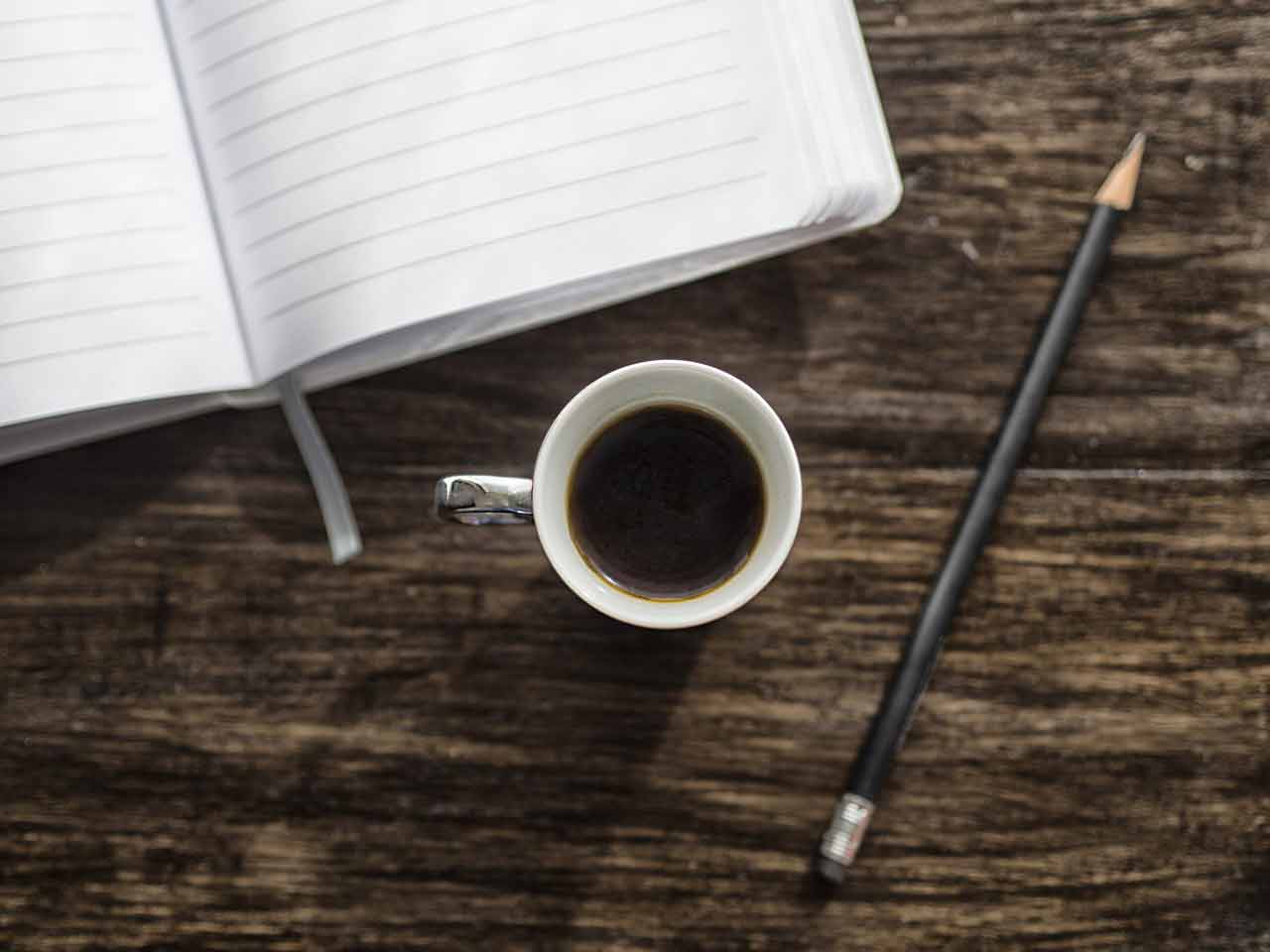 A photo of an empty journal, a cup of coffee, and a pencil.